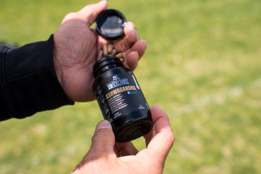 Person holding a black supplement bottle labeled 'Ashwagandha' against a green outdoor background